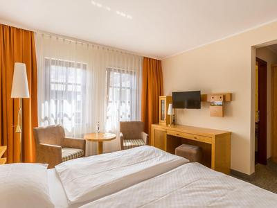 Helles Hotelzimmer mit Doppelbett, zwei Sesseln und orangefarbenen Vorhängen.