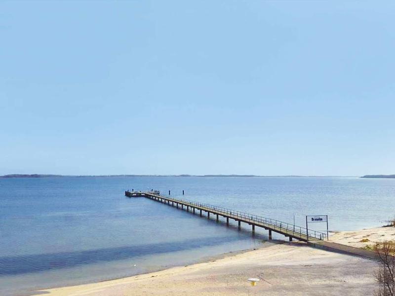 Long pier extends over sandy beach into calm blue sea under clear sky.