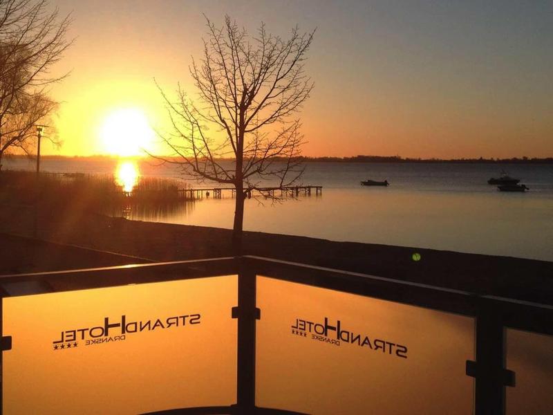 Sunset over calm water with a solitary tree and hotel railing in the foreground.