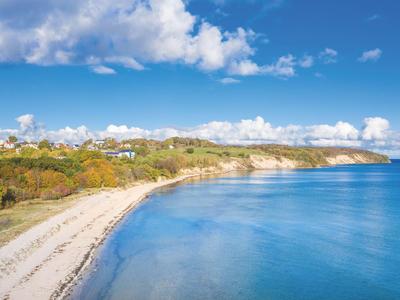 Küstenlandschaft mit Sandstrand, klarem blauen Wasser und bewaldeten Hügeln unter blauem Himmel.