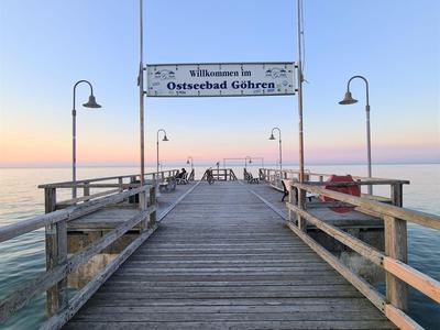 Langer Holzsteg führt über ruhiges Wasser bei Sonnenuntergang, Willkommensschild für Ostseebad Göhren.
