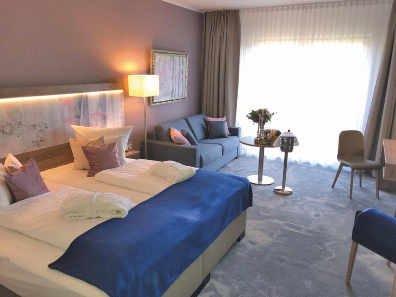 Bright hotel room with double bed, blue bedspread, sofa, and seating area by the window.