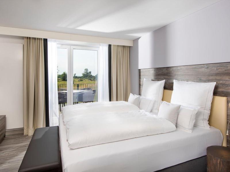 Helles Doppelzimmer mit großem Bett, TV, Balkon mit Glasgeländer und neutraler Einrichtung.