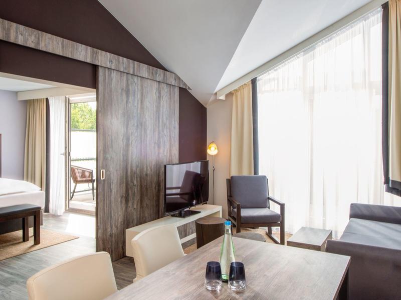 Modernes Hotelzimmer mit Holzakzenten, Sitzecke, Essbereich und Balkon mit natürlichem Licht.