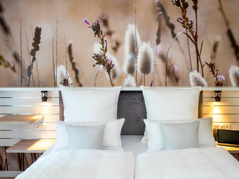 Modernes Schlafzimmer mit zwei Einzelbetten, weißen Kissen und Wanddekoration aus Gräsern und Blüten.