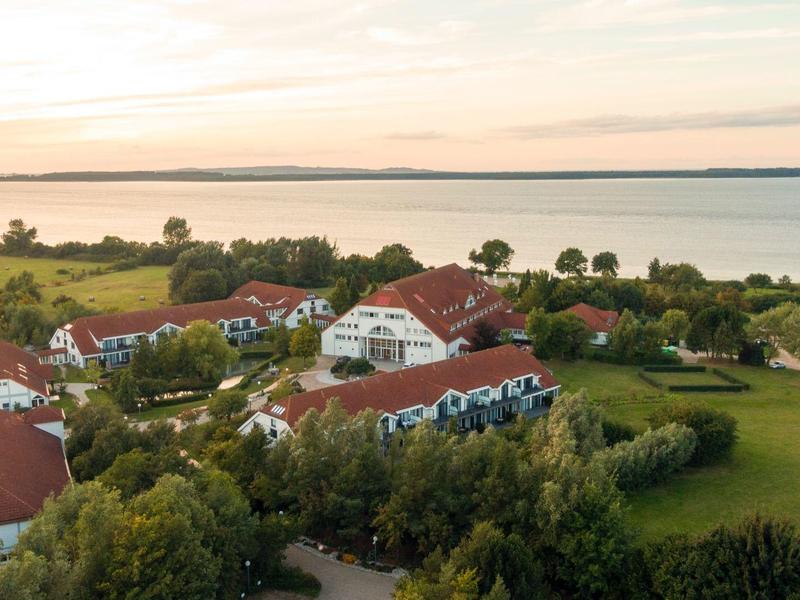 Landschaft mit grünen Feldern, Bäumen, Häusern und einem großen See im Hintergrund bei Sonnenuntergang.