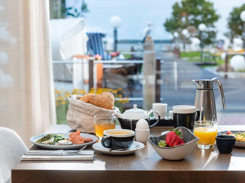 Frühstückstisch mit Brötchen, Kaffee, Orangensaft und frischem Obst vor Meerblick im Hintergrund.