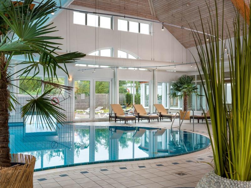 Innenraum mit großem Schwimmbad, Palmen und Liegestühlen vor bodentiefen Fenstern.