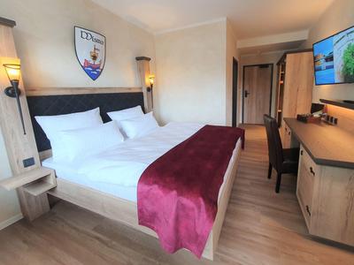 Hotelzimmer mit großem Doppelbett, weißen Bettwäsche, roter Decke und Holzmöbeln, Fernseher an der Wand.