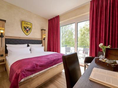 Helles Schlafzimmer mit großem Bett, rotem Überwurf und schweren roten Vorhängen am Fenster.