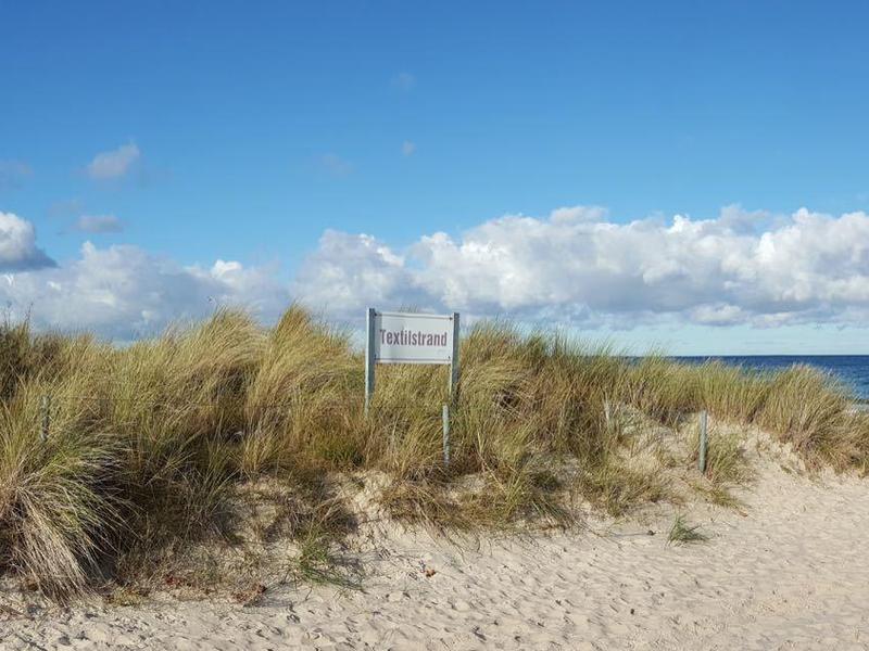 Strand mit Sand, Dünen und Gras, im Hintergrund Meer und blauer Himmel mit Wolken.