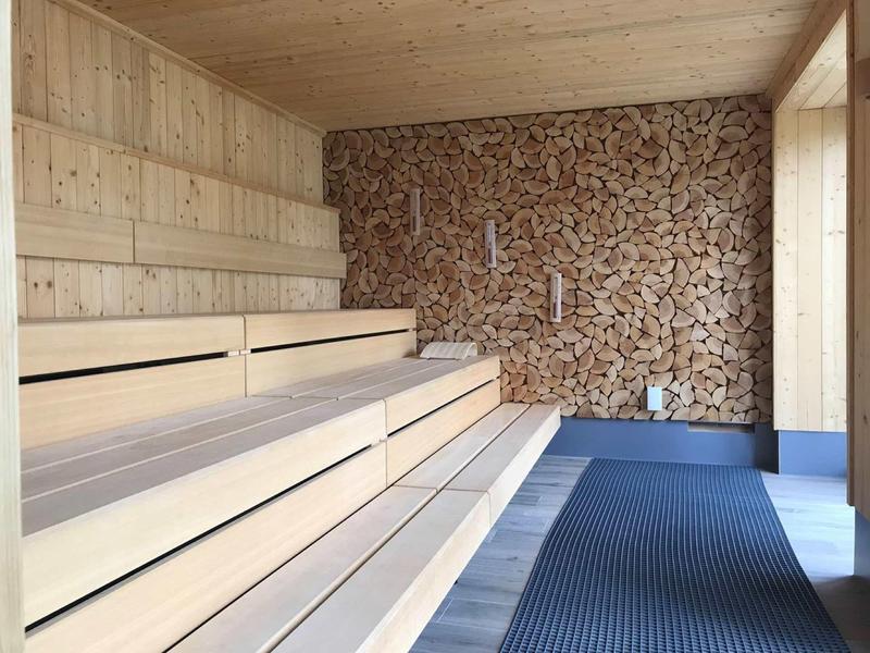 Sauna moderna con panche in legno chiaro e parete decorata con legna impilata.