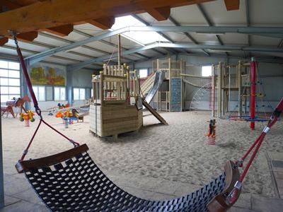 Aire de jeux intérieure avec sable, structure d'escalade, balançoire et hamac sous une verrière.