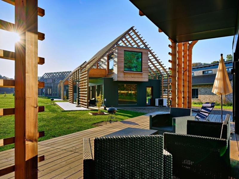 Modernes Holzhaus mit Glaswänden, Terrasse, Gartenmöbeln und sonnigem, grünem Außenbereich.