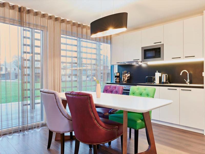 Moderner Küchenbereich mit weißem Schrank, bunter Stuhlreihe und großen Fenstern mit Vorhängen.