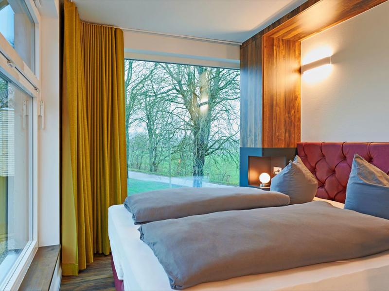 Helles Schlafzimmer mit großen Fenstern, gelben Vorhängen, rotem Polsterbett und grauer Bettwäsche.