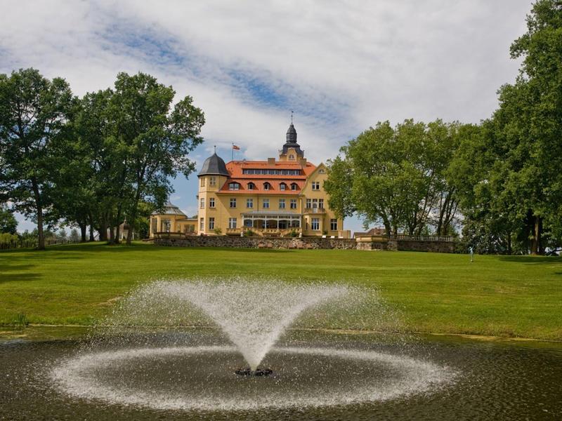 Schloss mit rotem Dach hinter großer Springbrunnenpfütze, umgeben von grünen Bäumen und Wiesen.