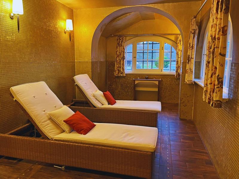 Gemütlicher Raum mit zwei weißen Liegen, warmem Licht und Fenster mit bunten Vorhängen.