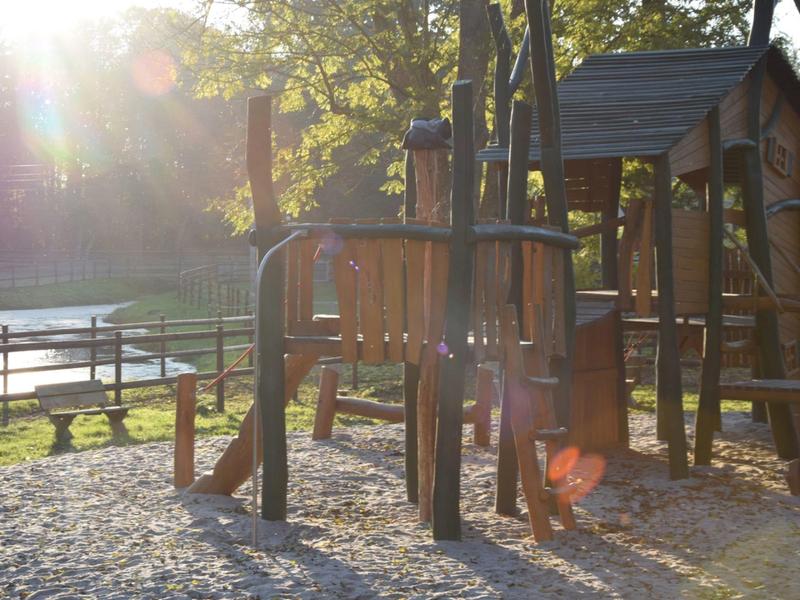 Kinderspielplatz mit Klettergerüst aus Holz, Rutsche, Sandboden und Sonnenlicht im Hintergrund.