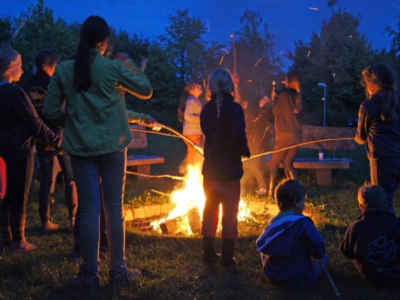 Menschen stehen und sitzen abends um ein Lagerfeuer im Freien, umgeben von Bäumen.