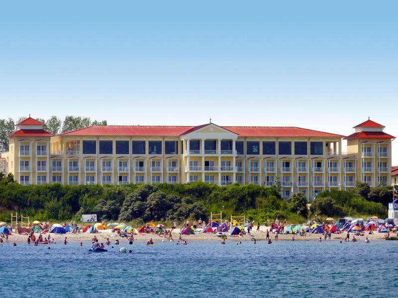 Großes Hotelgebäude mit roten Dächern an einem belebten Strand mit vielen Badenden.