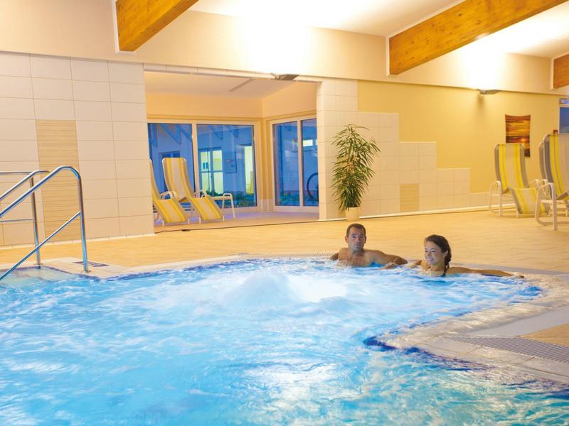 Zwei Personen entspannen im Whirlpool eines Innenpools mit Liegestühlen und Pflanzen.