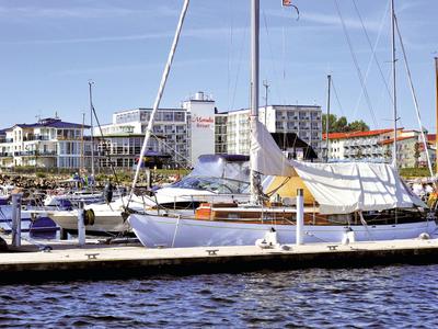 Segelboote liegen an einem Steg vor einem Hotel am Wasser unter blauem Himmel.
