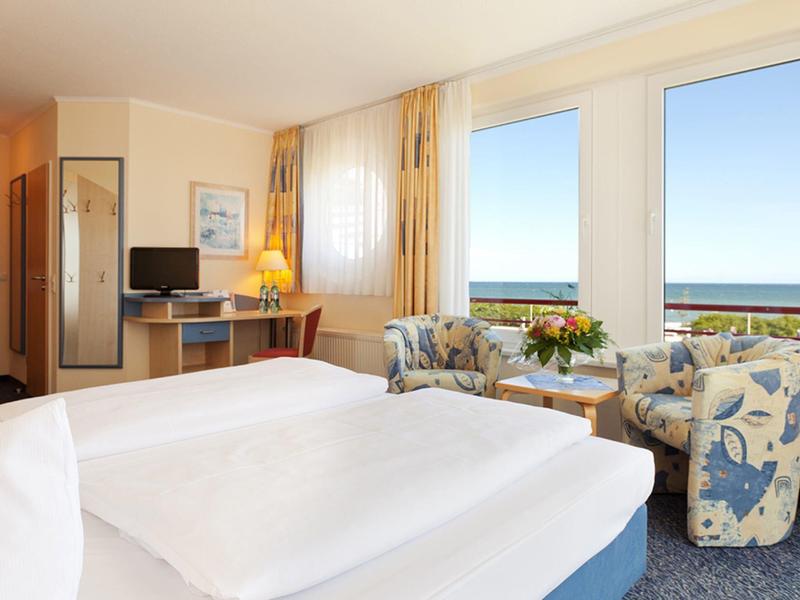 Hell eingerichtetes Hotelzimmer mit Doppelbett, Sitzbereich und Meerblick durch große Fenster.