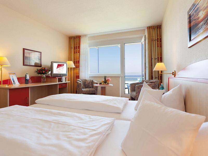 Helles Hotelzimmer mit zwei Betten, Sesseln und großem Fenster mit Blick aufs Meer.