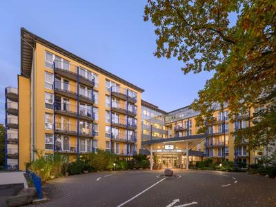 Modernes Hotelgebäude mit mehreren Etagen und Balkonen, umgeben von Bäumen bei Abendlicht.