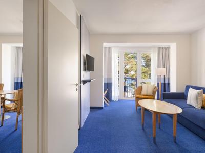 Helle Hotelzimmer mit blauem Teppich, Sofa, Tisch und Balkon mit Blick nach draußen.