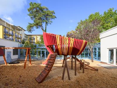 Kinderspielplatz mit Holzschiff und Rutschen in einem Hotelinnenhof bei klarem Himmel.
