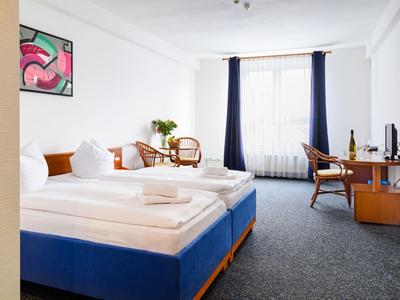 Bright hotel room with two single beds, desk, chairs, and large windows with curtains.