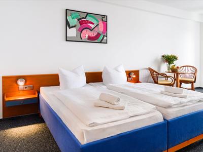 Two single beds with white bedding and towels in a bright hotel room with artwork and seating area.