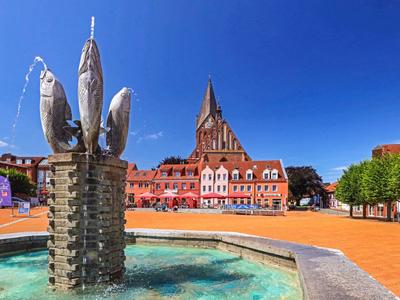 Brunnen mit Fischskulpturen vor einem roten historischen Gebäude bei klarem Himmel.