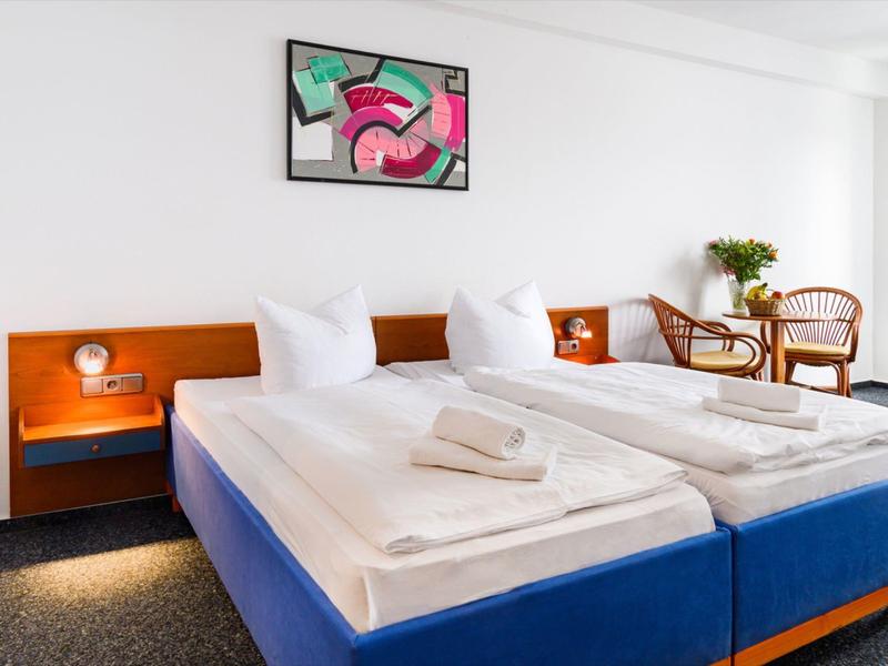 Helles Hotelzimmer mit zwei Betten, Nachttischen, Stühlen und moderner Wandkunst.