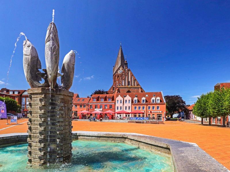 Brunnen mit Fischskulpturen vor einem roten historischen Gebäude bei klarem Himmel.