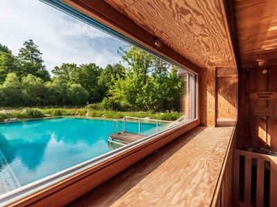Vista a través de una gran ventana hacia una piscina y árboles desde una casa de madera.