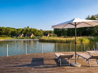 Steg mit Liegestühlen und Sonnenschirm am See vor Wald und Hütten bei klarem Himmel.