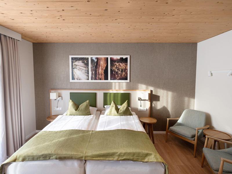 Habitación de hotel luminosa con dos camas, cojines verdes y área de asientos con sillones.