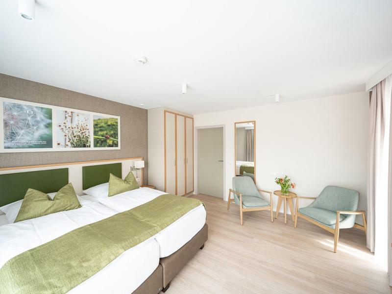 Habitación de hotel luminosa con dos camas, detalles verdes, área de estar y ventana con cortinas.