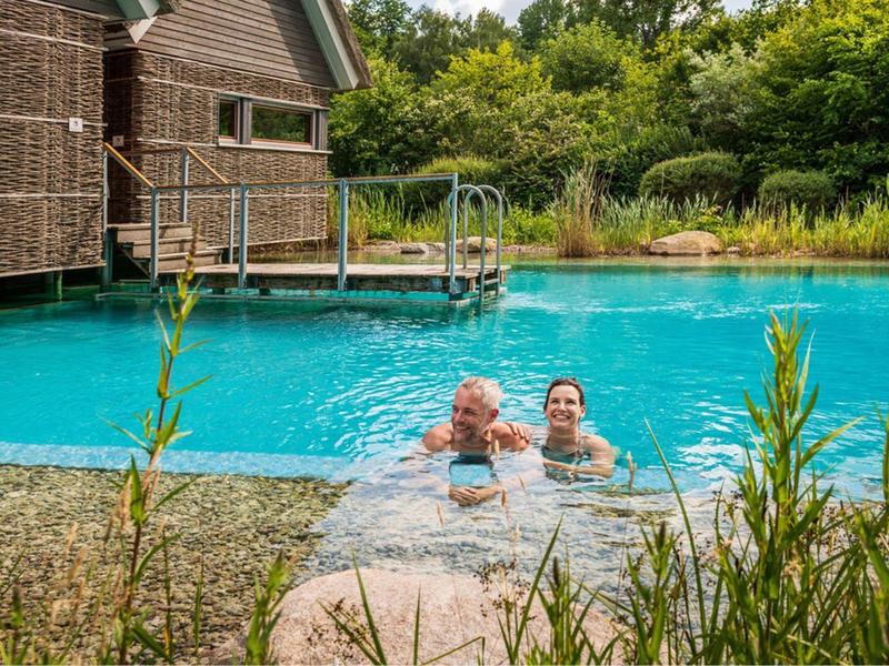 Dos personas nadan en un estanque natural junto a una casa de madera en entorno verde.