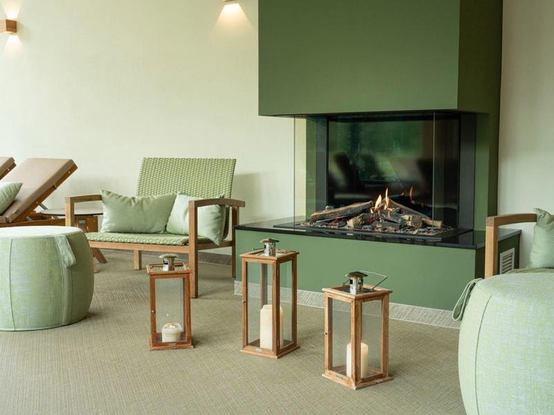 Área de estar acogedora en hotel con sillones verdes, chimenea de madera y faroles en el suelo.