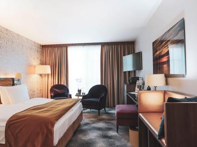 Modern hotel room with double bed, two armchairs, and wall-mounted TV.