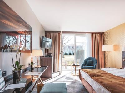 Modern furnished hotel room with bed, chairs, desk, and balcony in daylight.