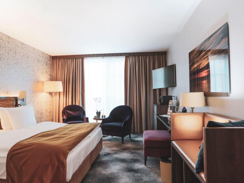 Nowoczesny pokój hotelowy z dużym łóżkiem, dwoma fotelami i telewizorem z płaskim ekranem przy oknie.