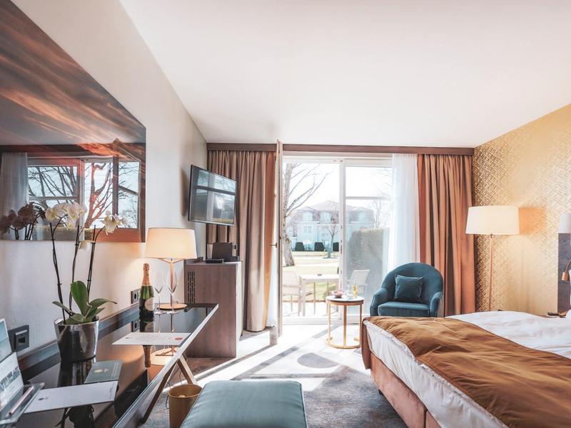 Nowoczesny pokój hotelowy z dużym łóżkiem, fotelami i balkonem z widokiem na zieleń.