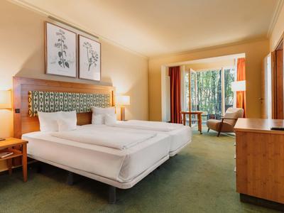 Helles Hotelzimmer mit zwei Betten, Holzmöbeln und Zugang zu einem Balkon mit Waldblick.