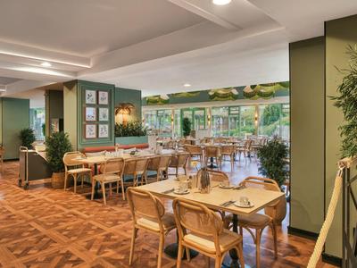 Ristorante moderno con pavimenti in legno, tavoli e sedie in legno, grandi finestre e piante verdi.