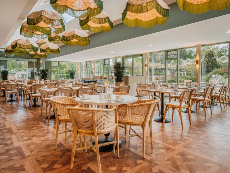 Helles Restaurant mit vielen Holztischen und Stühlen, großer Fensterfront und dekorativen Lampen.